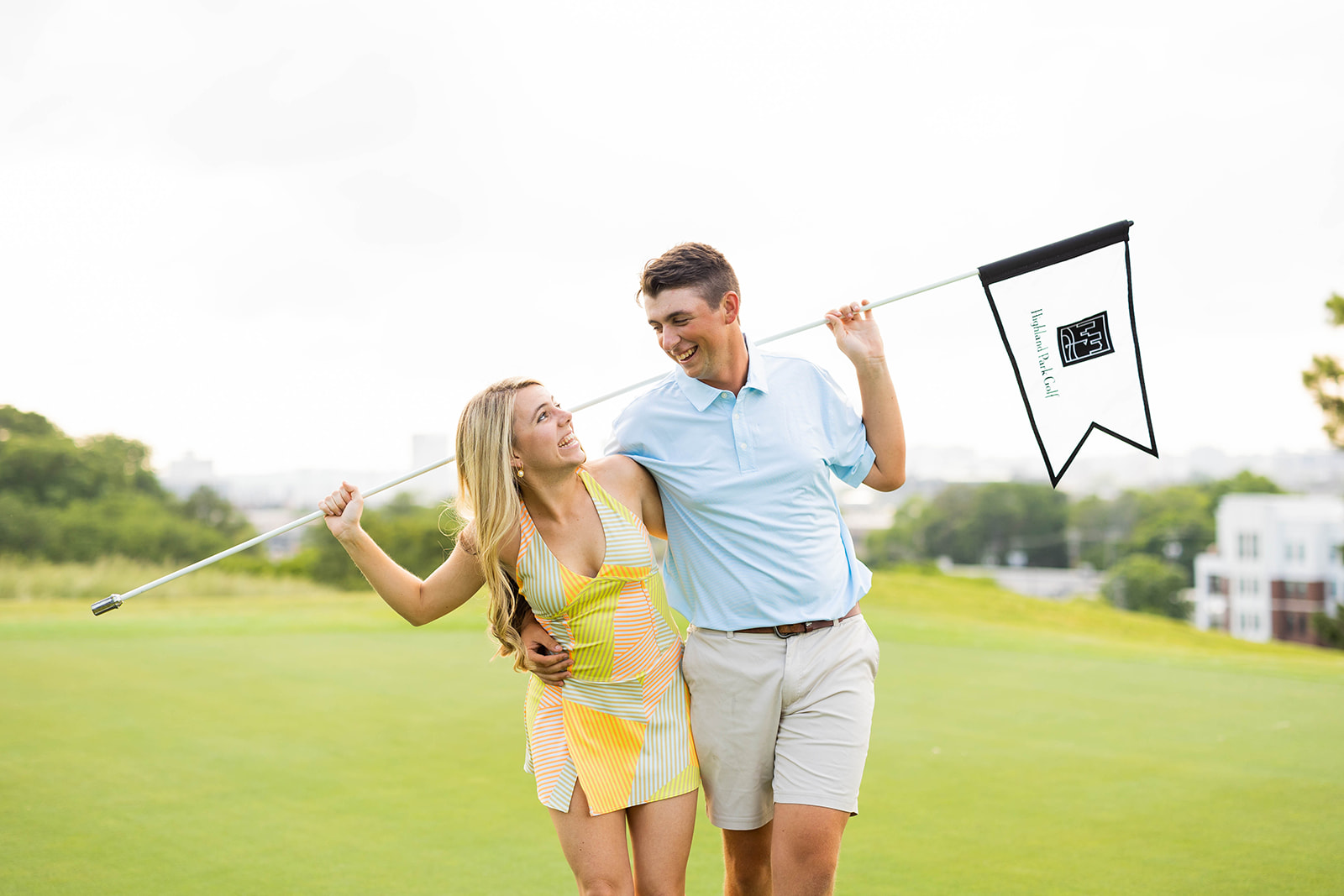 An engaged couples is standing on a golf course. They are both holding the hole flag over their shoulders and smiling at each other. 