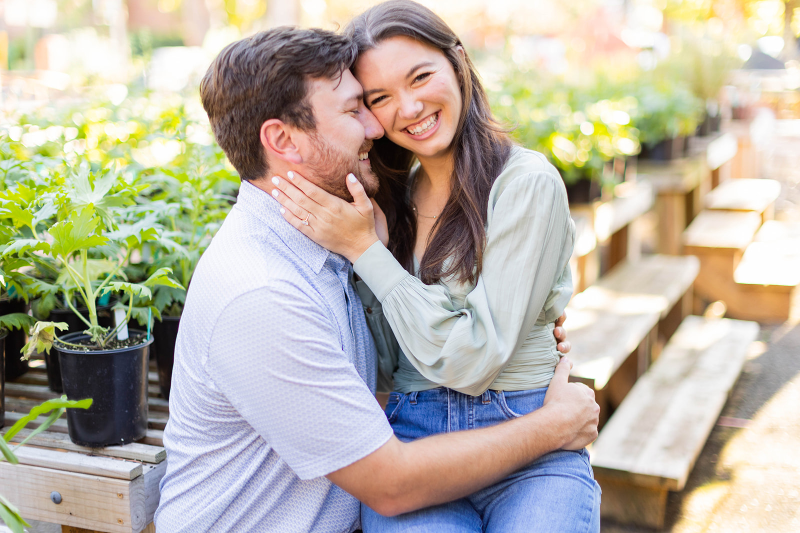 An engaged couple sits together at a plant nursery, embracing and smiling at the camera. 