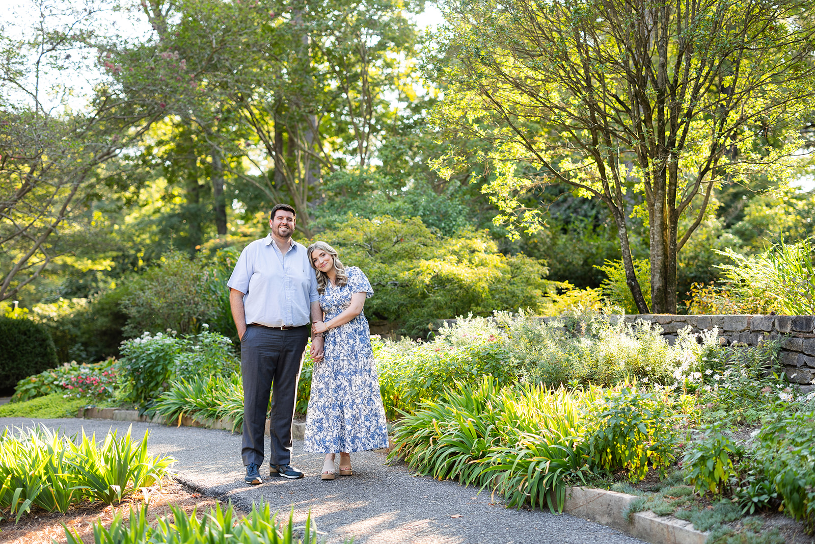 An engaged couple stands together on the sidewalk at the Birmingham Botanical Gardens. The bride holds on to her fiancé's arm and they both smile at the camera. 