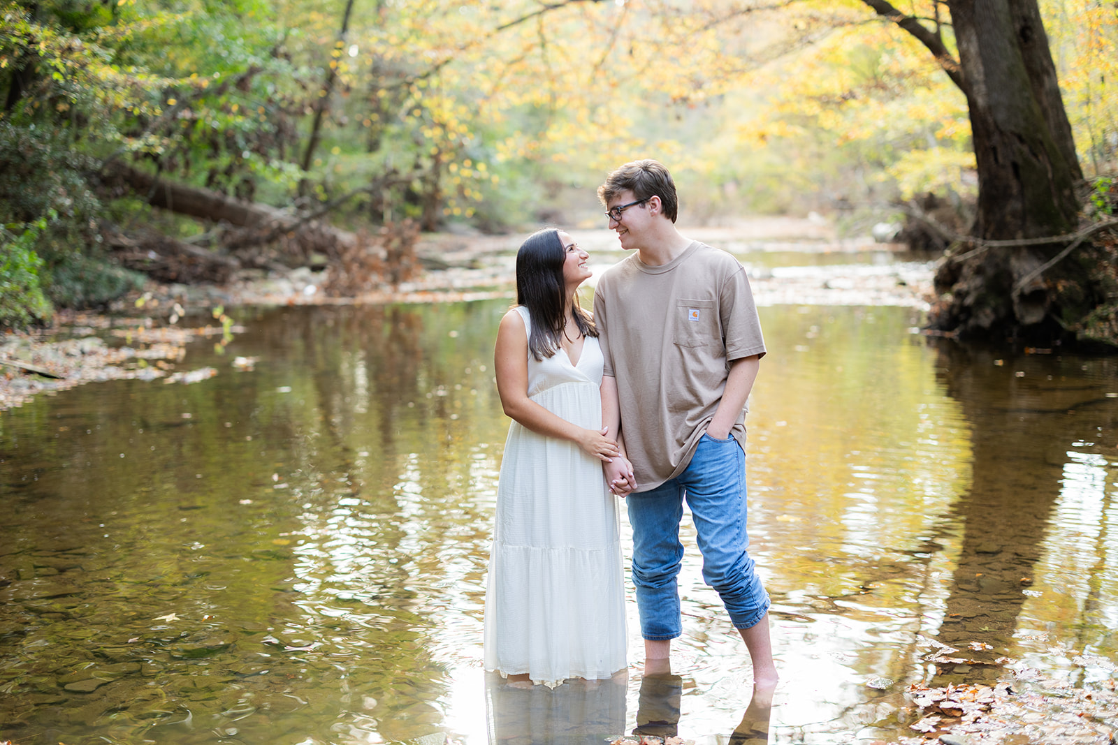 An engaged couple stands in a shallow river at Oak Mountain State Park in Birmingham, AL. They hold hands and smile at each other. 