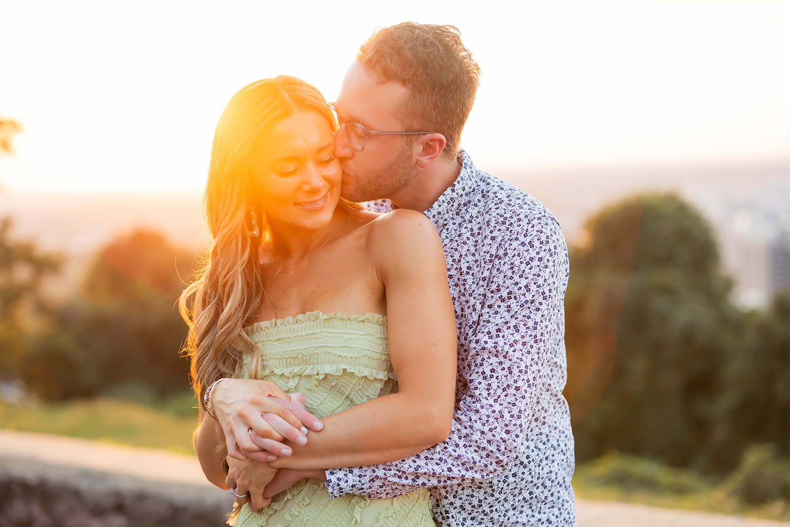 An engaged couple stands together at a city overlook at sunset. The groom embraces his bride from behind and kisses her cheek. 