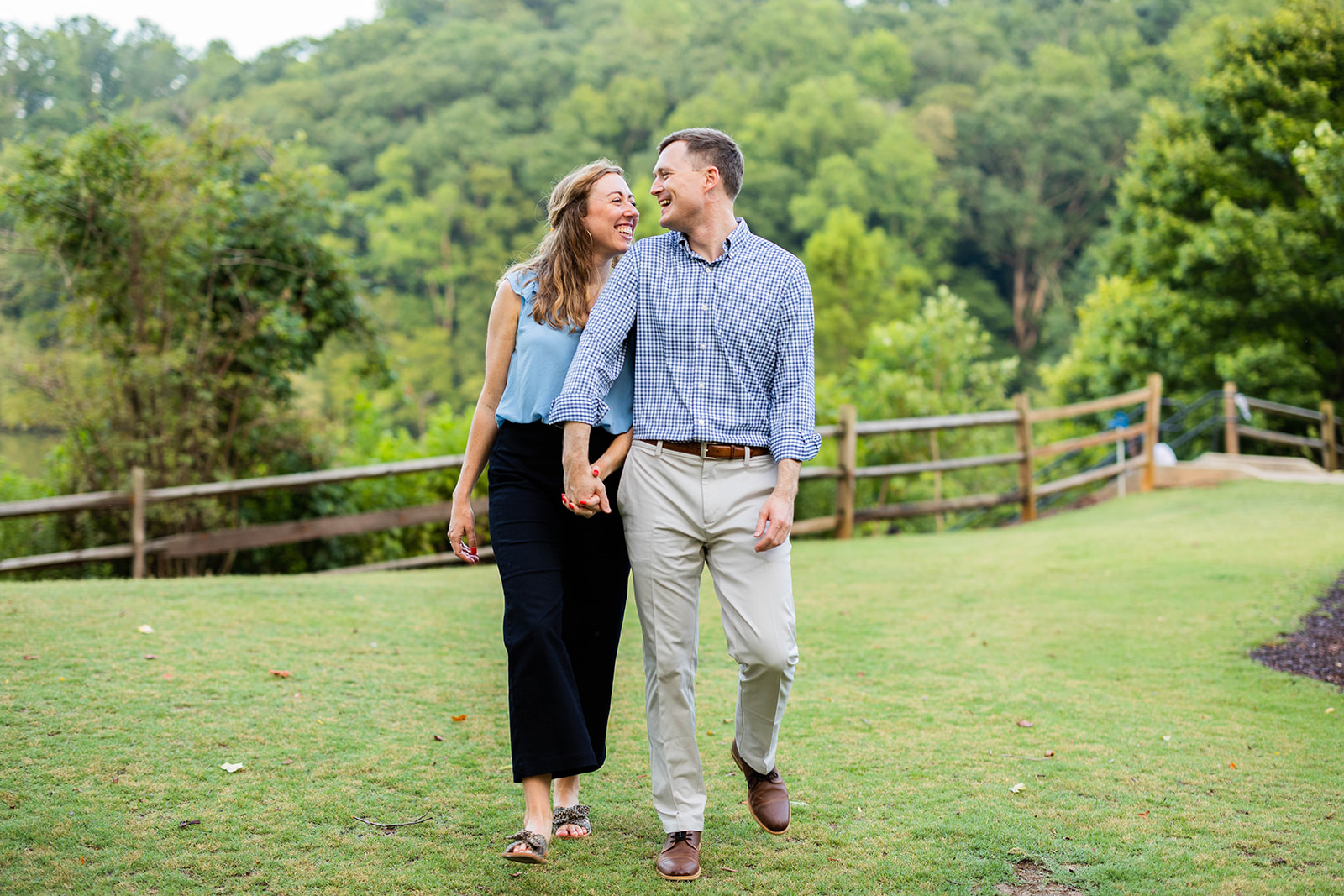 An engaged couple walks through a field of grass, holding hands and laughing with each other. 