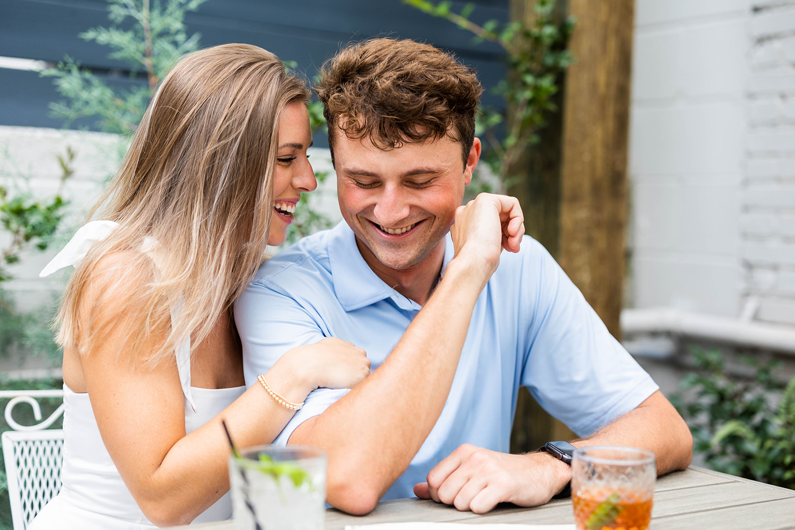 An engaged couple sits at a table at an outdoor patio, sharing drinks and laughing with each other. 