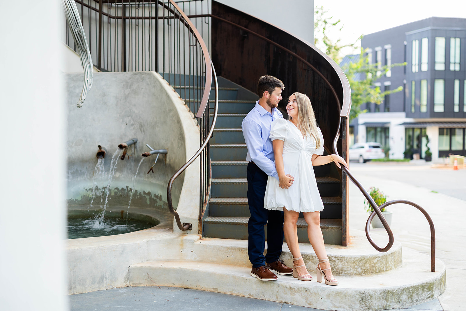 An engaged couple poses on a set of stairs together. They are holding hands and smiling at each other. 