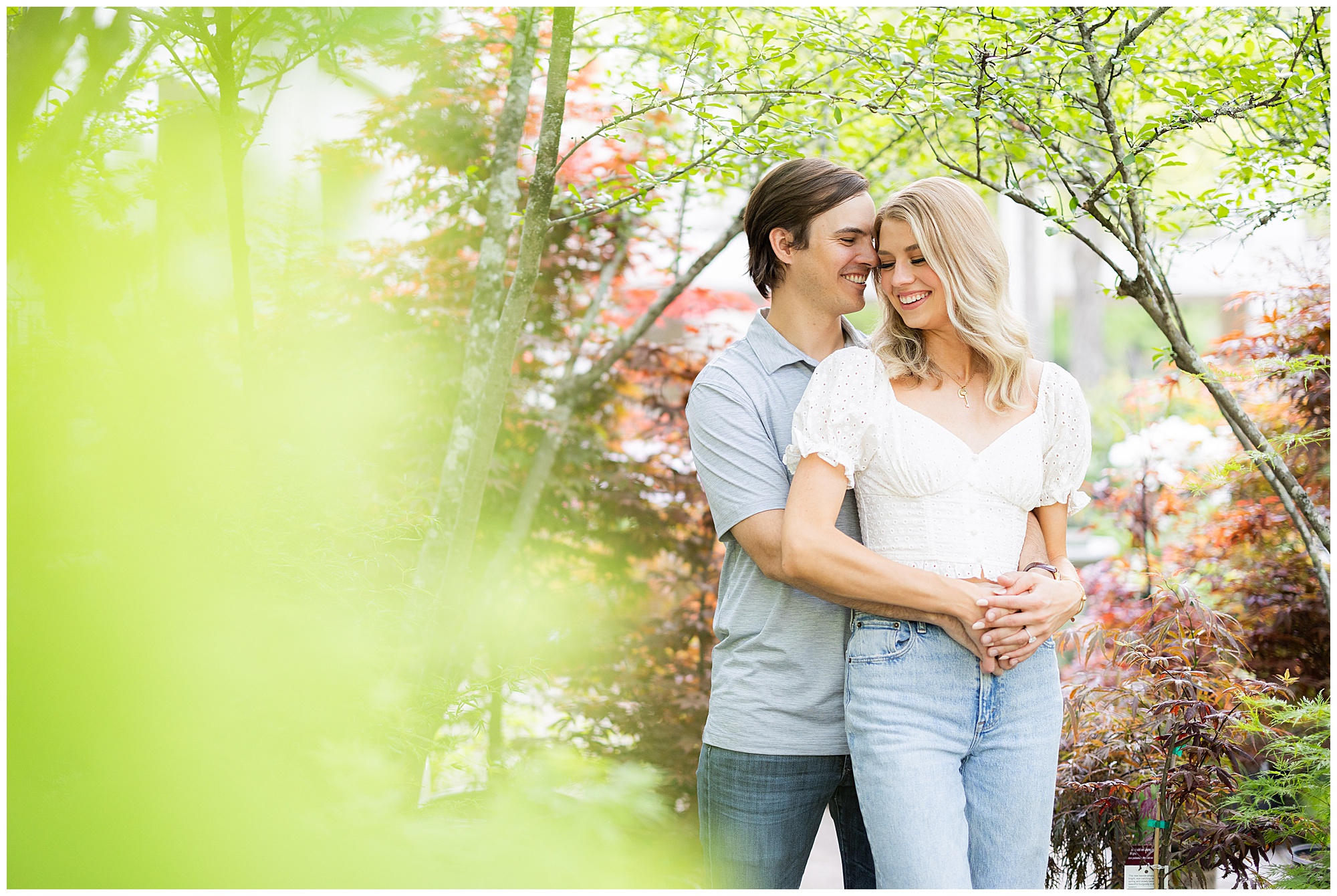 An engaged couple stands amidst the trees at the Birmingham Botanical Gardens. They embrace each other and smile. 