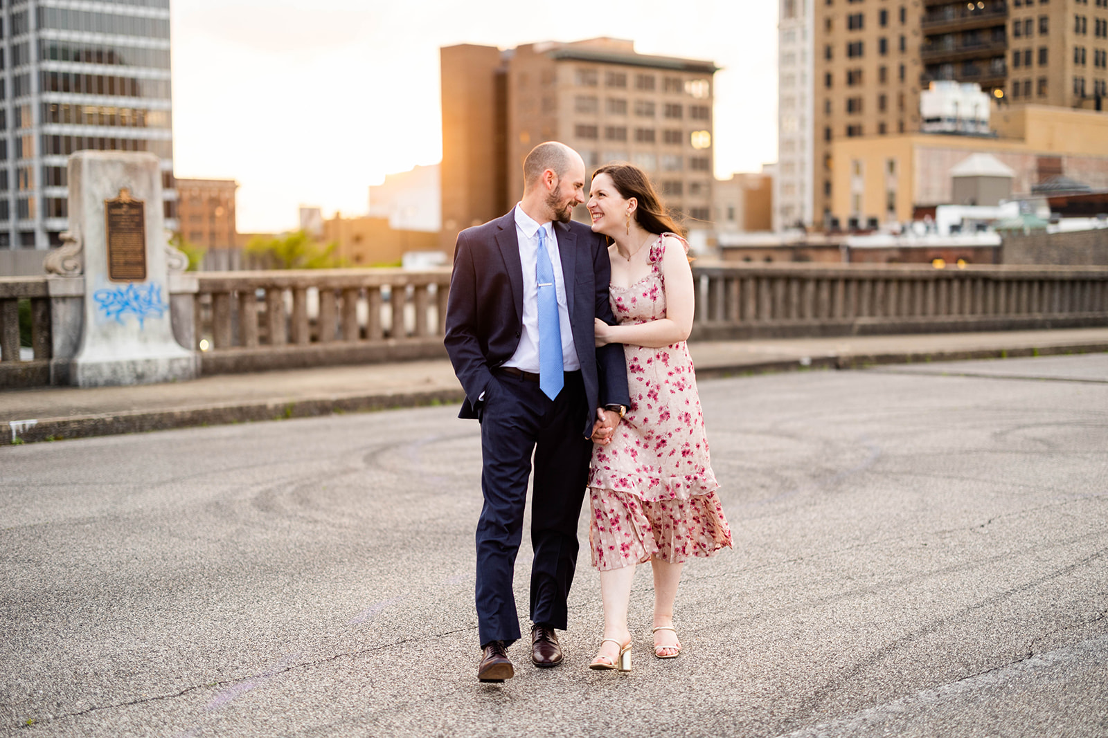 An engaged couple poses on the Pedestrian Bridge in Birmingham, AL. The bride is holding the arm of her fiancé and they're looking at each other and smiling. 