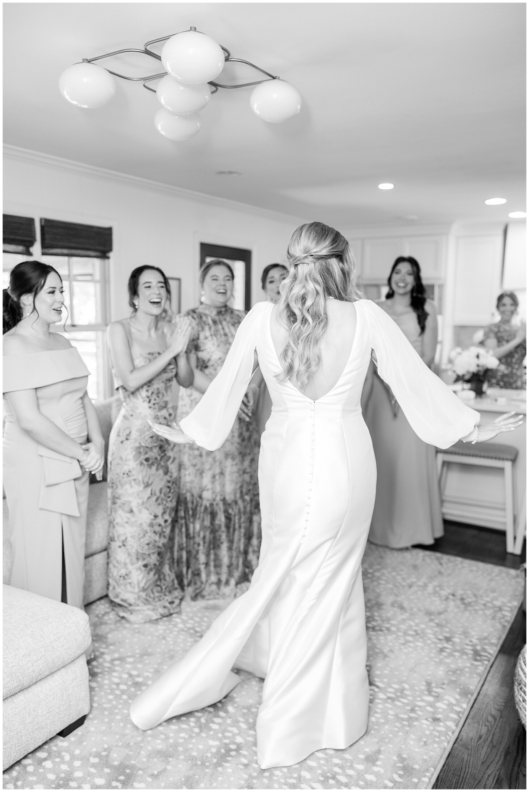 A black and white image of Faith showing all her bridesmaids what she looks like in her wedding dress. Her bridesmaids are smiling and cheering for her. 