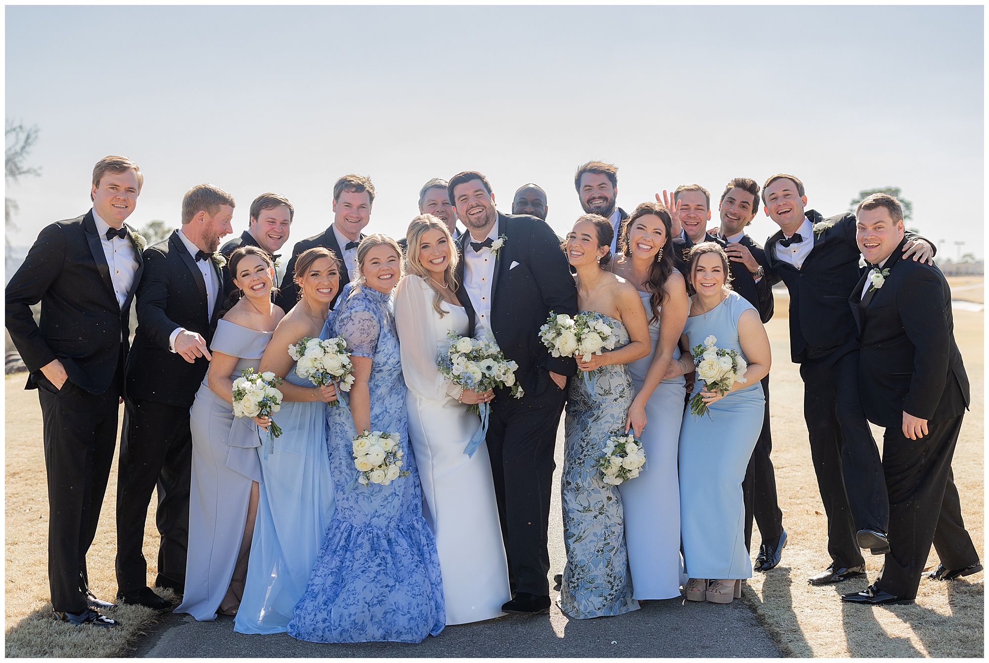 Faith and James are surrounded by their bridesmaids and groomsmen. The girls are wearing various blue dresses and the guys are wearing black tuxedos. 