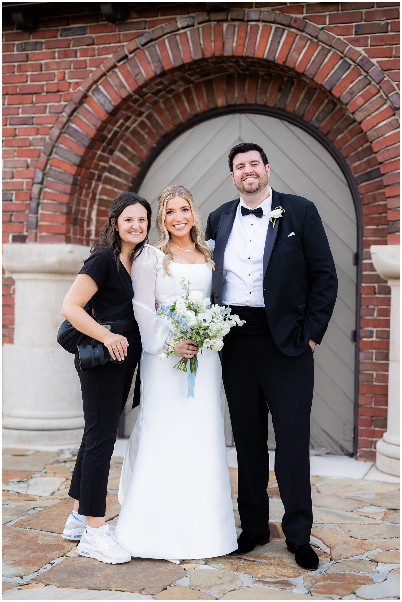 Eleanor, the wedding photographer, stands and smiles next to Faith and James. 