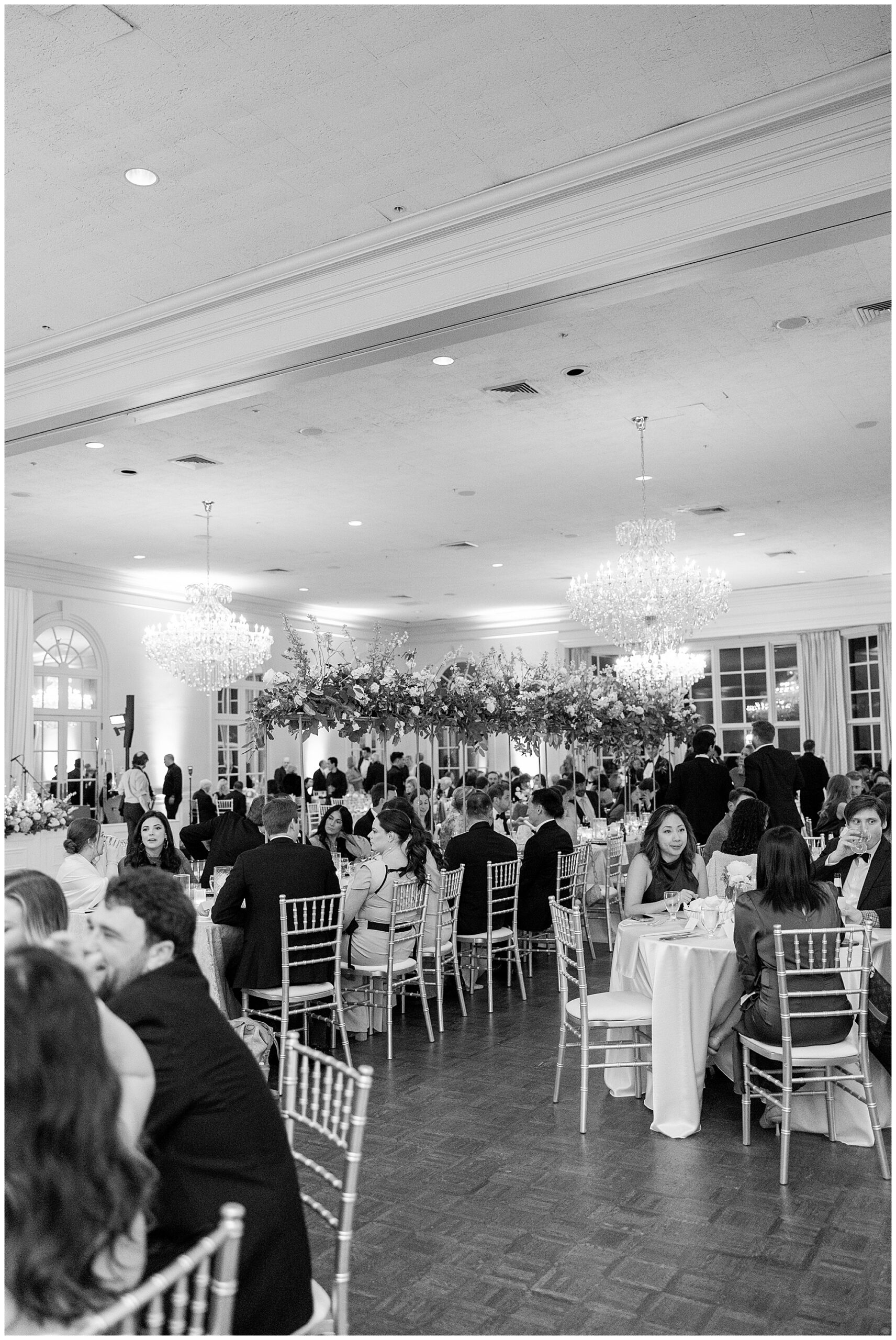 A black and white wide shot of the room as guests eat their dinner and mingle. 