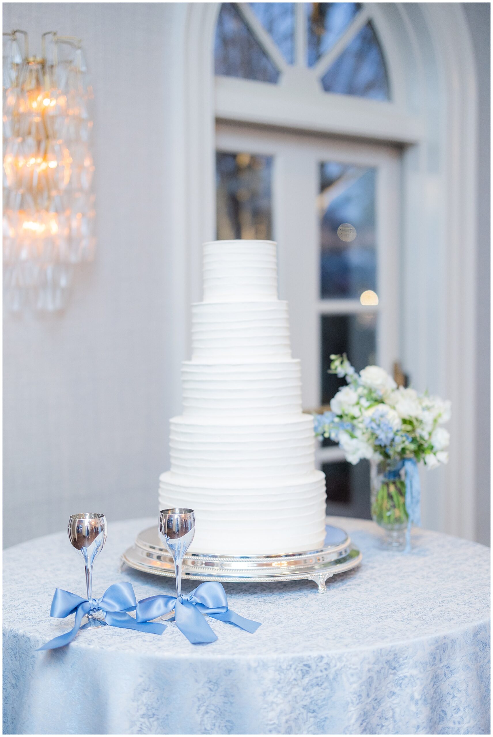 A 5 tier white wedding cake sits on a table next to 2 chalices and Faith's bridal bouquet. 