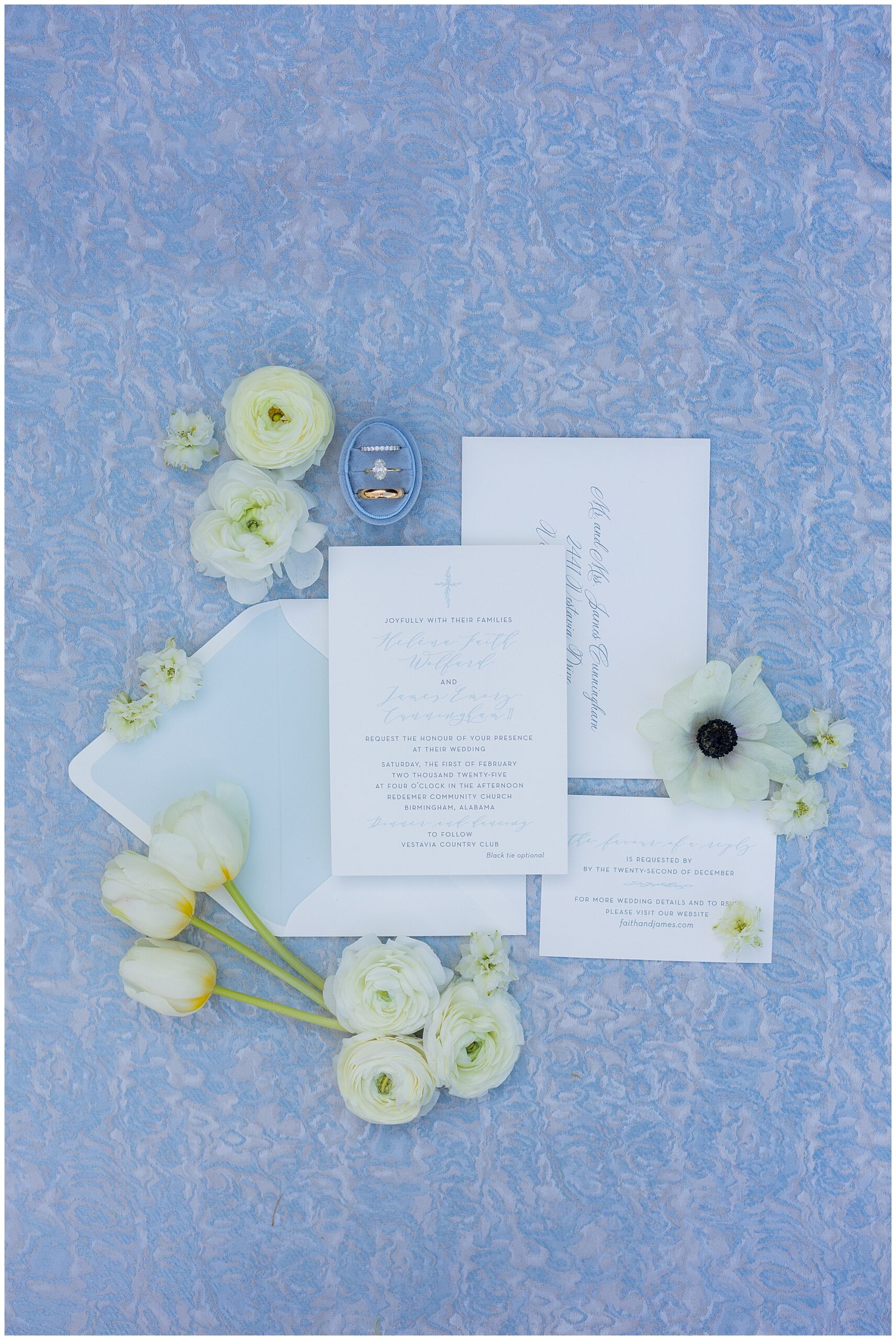 Faith and James' invitation suite, wedding rings and styling blooms are laid out on a blue and silver linen. 